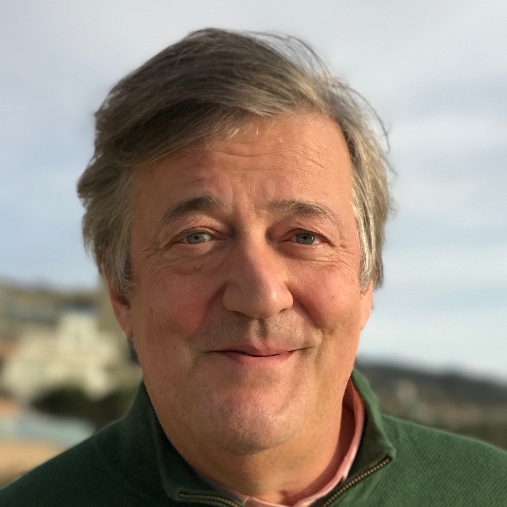 Stephen Fry smiling at the camera with blue sky and white cliffs in the background.