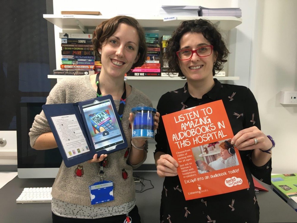 Two women looking at the camera and holding up a tablet and an orange poster.