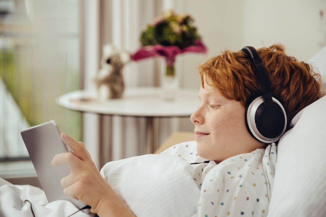 Boy listening to books in hospital bed wearing headphones and looking at a tablet.