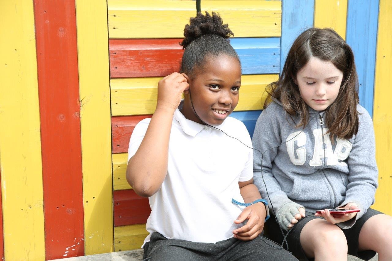 Two children sitting in front of a yellow, red and blue striped wall sharing headphones and looking at an iPod.