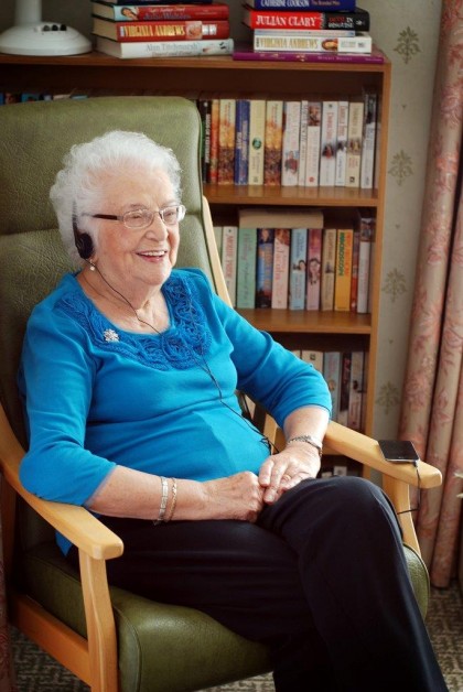 Older woman sat in a chair in front of a bookcase, wearing headphones.