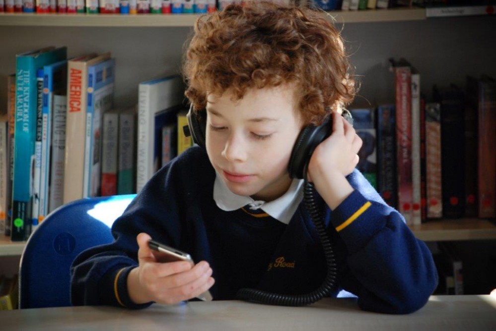 Boy in school uniform sitting in front of a bookshelf, listening to an audiobook through headphones.