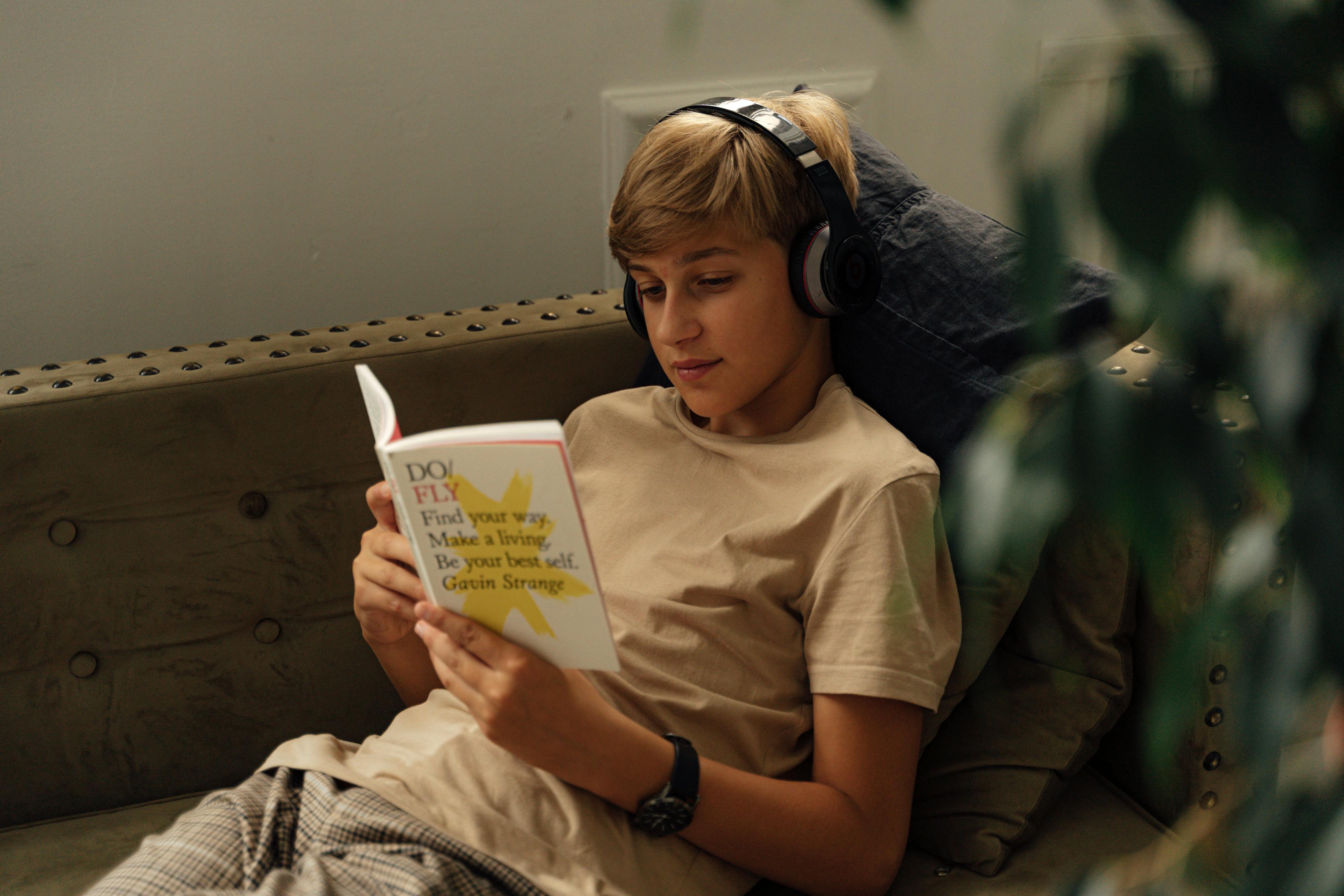 A young teen boy sitting on a sofa holding a book with headphones on